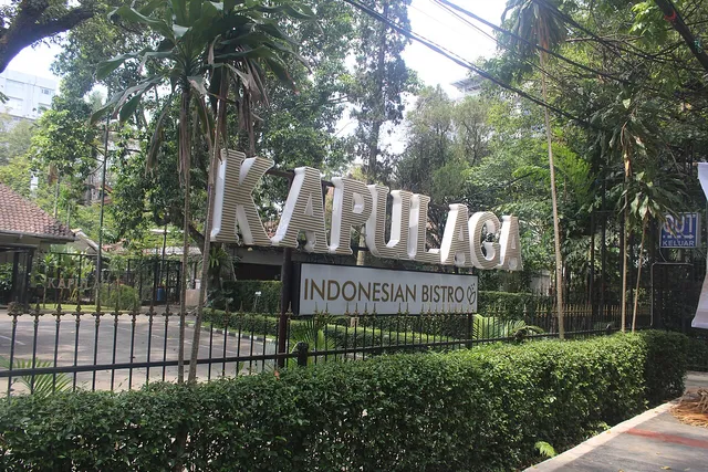 Penanda Kapulaga Indonesian Bistro di depan pepohonan dan pagar, Oktober 2024. / Kapulaga Indonesian Bistro signage in front of trees and a fence, October 2024.