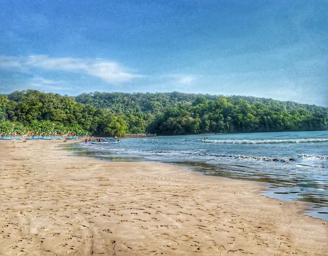 ID: Pesisir Pantai Pangandaran dengan bukit hijau dan aktivitas perahu. EN: Pangandaran shoreline, forested hills, and boats near the beach.