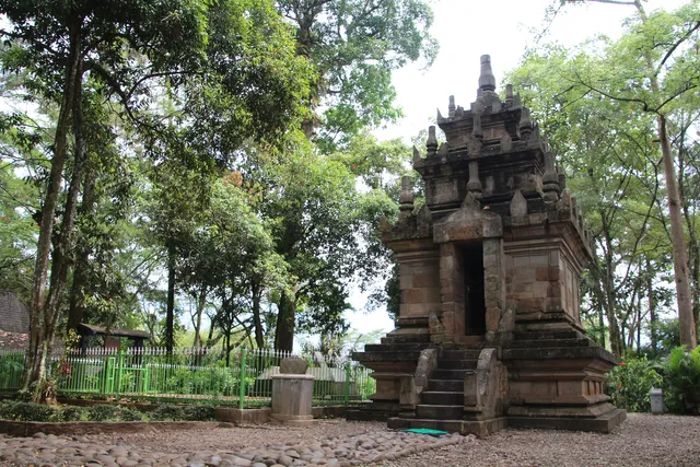 ID: Candi Cangkuang dari depan dengan pepohonan dan pagar di sekitarnya. EN: The facade of Cangkuang Temple with surrounding trees and fencing.