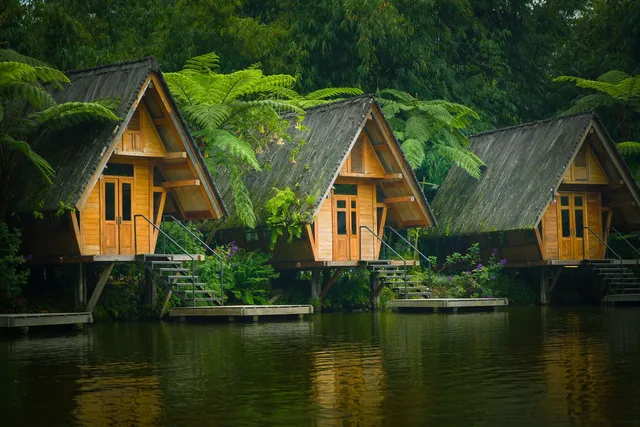 ID: Tiga saung kayu beratap pelana dengan tangga menuju danau, di antara pakis dan bambu di Dusun Bambu. EN: Three gabled wooden pavilions with steps down to the lake, among ferns and bamboo at Dusun Bambu.