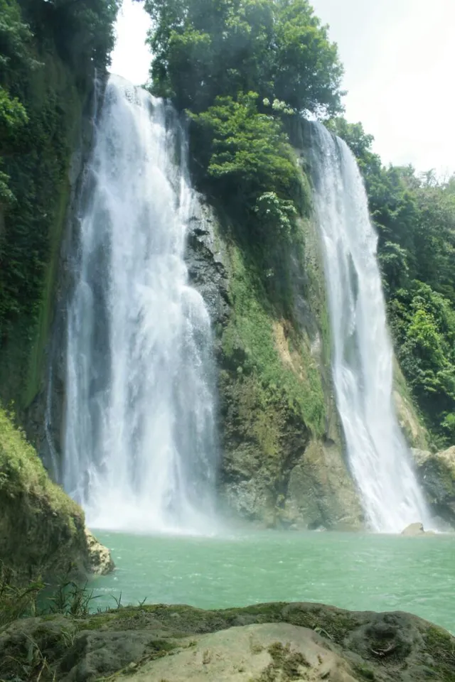 Dua aliran Curug Cikaso jatuh ke kolam di bawah tebing hijau. / Two flows at Curug Cikaso drop into a pool beneath green cliffs.