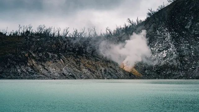 ID: Danau Kawah Putih dengan lereng berbatu dan kepulan uap di seberangnya. EN: Kawah Putih lake with rocky slopes and a plume of steam on the far shore.