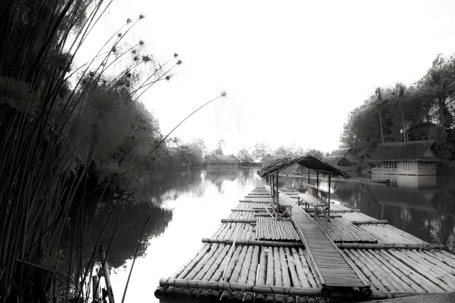 ID: Foto hitam putih rakit bambu beratap di tepi danau Kampung Sampireun. EN: A monochrome photograph of a roofed bamboo raft beside Kampung Sampireun’s lake.