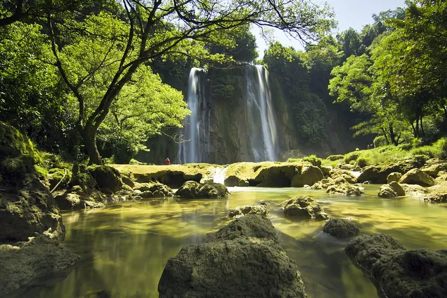 Curug Cikaso dengan bebatuan dan aliran air di depan; seorang pengunjung sangat jauh, wajah tidak dikenali. / Curug Cikaso with foreground rocks and water; a distant visitor has no identifiable face.