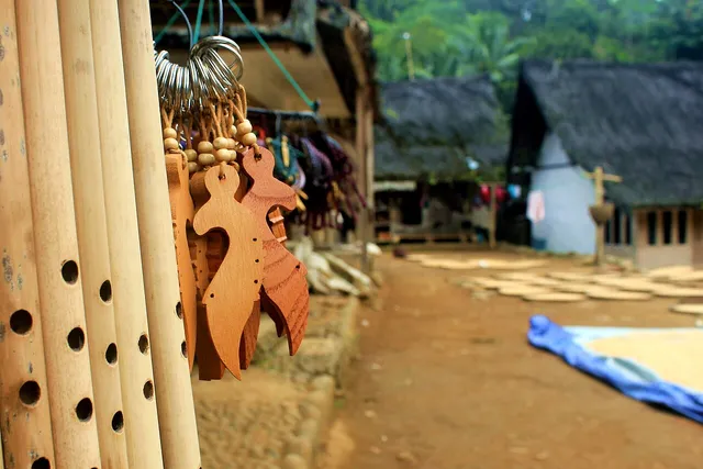 ID: Suling dan gantungan kunci berbentuk kujang, dengan rumah Kampung Naga di latar belakang. EN: Flutes and kujang-shaped keyrings, with Kampung Naga houses in the background.