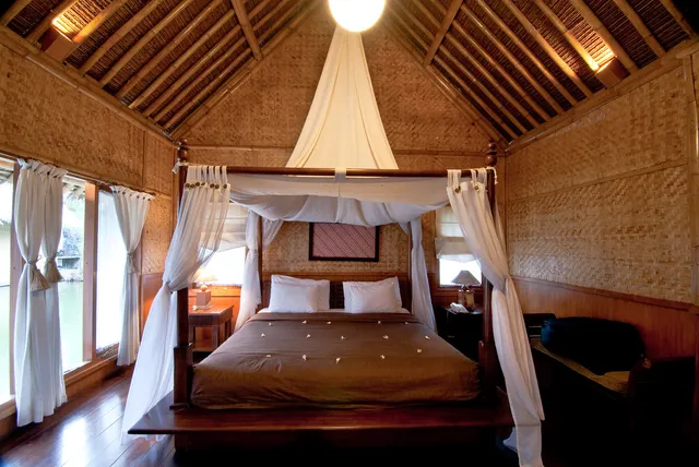 ID: Tempat tidur berkelambu dengan dinding anyaman bambu dan jendela menghadap danau di Kampung Sampireun. EN: A canopy bed, woven bamboo walls and a lake-facing window at Kampung Sampireun.