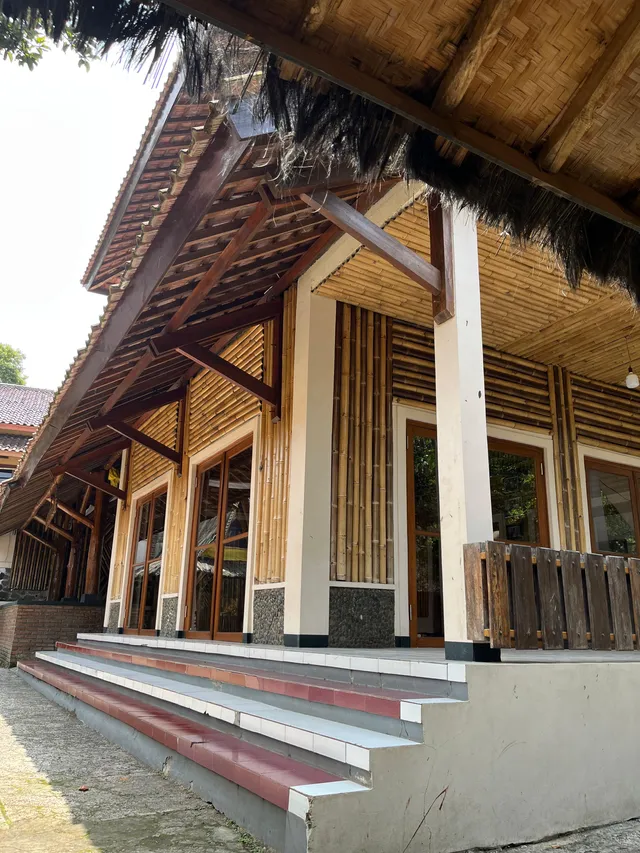 ID: Tampak samping bale dengan dinding bambu, tiang, dan tangga di Kampung Adat Cireundeu. EN: The side of a community hall with bamboo walls, columns and steps in Kampung Adat Cireundeu.