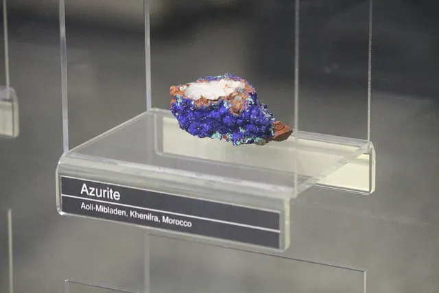 ID: Spesimen azurit berwarna biru pada dudukan transparan di Museum Geologi Bandung. EN: A blue azurite specimen on a transparent stand at Museum Geologi Bandung.