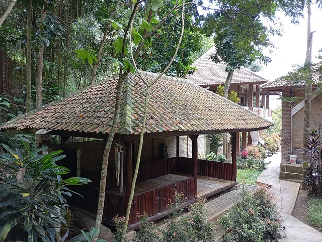 ID: Saung beratap genteng di antara pepohonan dan jalan setapak di Kampung Adat Cireundeu. EN: A tiled-roof pavilion among trees and a footpath in Kampung Adat Cireundeu.